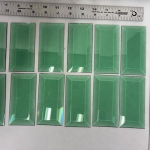 May include: A collection of twelve rectangular, seafoam green glass tiles. Each tile has a beveled edge and a glossy finish. Rulers are placed above and to the side of the tiles, showing measurements in inches. The tiles are arranged in three rows of four.