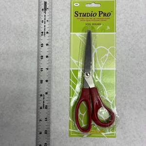 May include: Foil shears with red handles in a green and white package. The package reads "Studio Pro" and "Explore the art of stained glass with quality glass tools." A ruler is to the left of the shears.