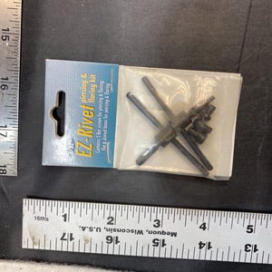 May include: A package labeled "EZ-Rivet piercing & flaring kit" containing T-bar screws and flat & domed bases. The kit is packaged in a clear plastic bag. A metal ruler is next to the package.