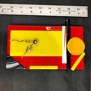 May include: A clock made of colorful glass tiles in red, yellow, and black. The clock face is yellow with black hands.
