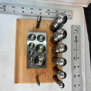 May include: A wooden jewelry-making block with a metal forming block and various metal tools. The forming block has multiple circular indentations. Metal tools of various sizes are arranged around the block. Rulers are also present.