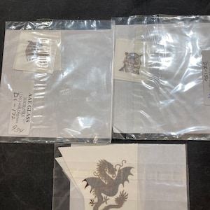 May include: Three clear plastic bags containing three different iron-on transfers. The transfers are of a dragon, a tiger, and a bird. The bags have labels with the text "AAE GLASS HIGH FIRE 1250-1400 Degrees DL-172 934", "DX-156", and "DC-175 937 1250-1400 Degrees".