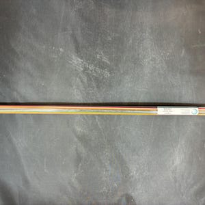 May include: A clear plastic tube containing multiple thin, colorful rods. The rods are a mix of orange, yellow, red, and other colors. A white label with text is visible on the tube. The tube is lying horizontally on a dark surface.