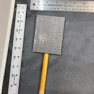 May include: A gray sanding block with a wooden handle. The sanding block is attached to the handle with a metal screw.