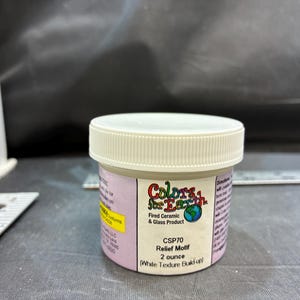 May include: A small, round, beige plastic jar with a white screw-top lid containing Colors for Earth CSP70 Relief Motif white texture build-up. The label indicates a 2 ounce volume. A metal ruler is visible in the background for scale.