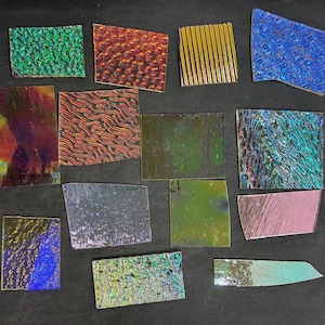 May include: Assortment of iridescent glass pieces in various shapes and textures. Colors include green, blue, red, and gold. Some pieces have textured surfaces, while others are smooth. A metal ruler is visible.