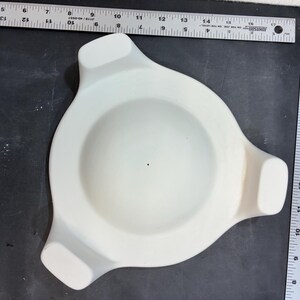May include: A white ceramic tripod bowl with a smooth, matte finish and a central dot. The bowl is positioned next to two metal rulers, showing a diameter of approximately 6 inches and a height of approximately 6 inches on the scale.