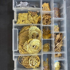 May include: A clear plastic organizer filled with various metal craft supplies. The compartments hold gold-colored filigree circles, floral shapes, and other decorative elements. There are also silver-colored metal pieces and small, brown, rounded beads. A metal ruler is at the top.