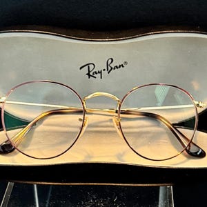 May include: Round eyeglasses with gold frames and pink rims rest inside an open case. The case is beige with a black border and the Ray-Ban logo. The glasses have a classic, stylish design.