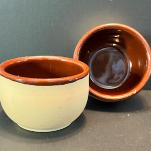 May include: Two small, handmade ceramic bowls. The exterior is a creamy beige, while the interior and rim are a rich brown. The bowls are round with a slightly flared rim. The bowls are likely for serving dips or small portions.