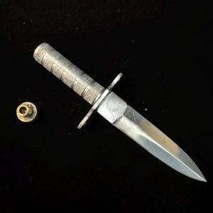 May include: A silver-toned metal dagger featuring a textured, cylindrical handle with a crossguard and a sharp, pointed blade. Beside the dagger lies a small, circular, brass-colored screw-on cap with a threaded metal post extending from its center.