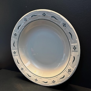 May include: A round, off-white ceramic soup bowl featuring a wide rim decorated with a repeating pattern of dark green four-petaled floral motifs and curved line accents. The bowl has a smooth, glossy finish and a slightly raised, textured border.