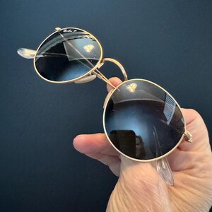 Pu&ograve; includere: Occhiali da sole con montatura dorata e lenti scure. Gli occhiali da sole hanno una classica forma rotonda e naselli trasparenti. Il logo Ray-Ban &egrave; visibile sulla lente superiore sinistra. Su sfondo scuro.