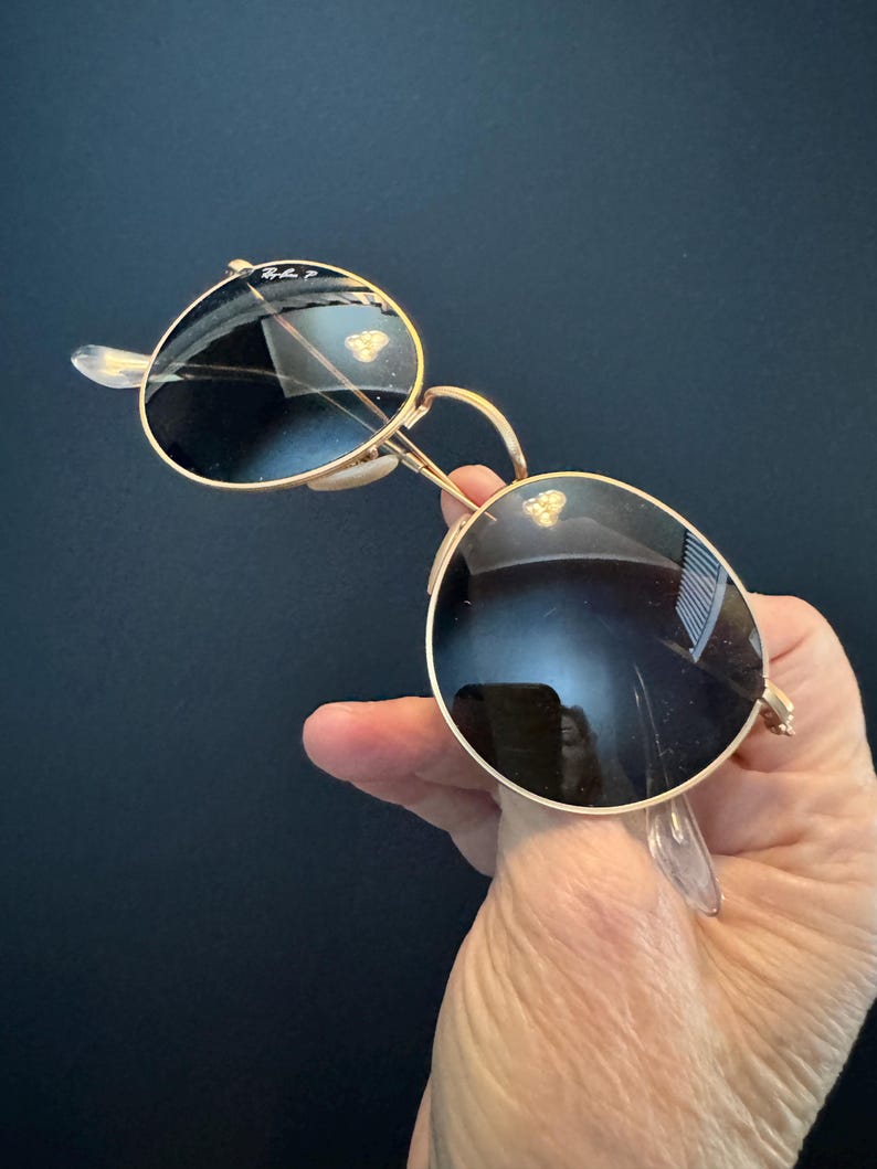 Pu&ograve; includere: Occhiali da sole con montatura dorata e lenti scure. Gli occhiali da sole hanno una classica forma rotonda e naselli trasparenti. Il logo Ray-Ban &egrave; visibile sulla lente superiore sinistra. Su sfondo scuro.