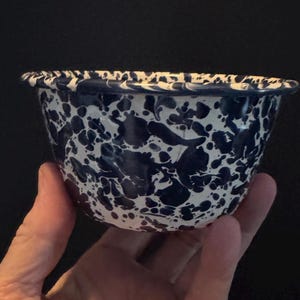 May include: A small, round bowl with a speckled pattern of navy blue and white. The bowl has a slightly flared rim and is held in a hand against a dark background. The bowl's design is reminiscent of enamelware.