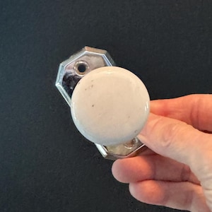 May include: A vintage door knob with a white speckled ceramic handle and a shiny silver-colored metal base. The knob is round and held by a hand against a dark background. The base is octagonal.