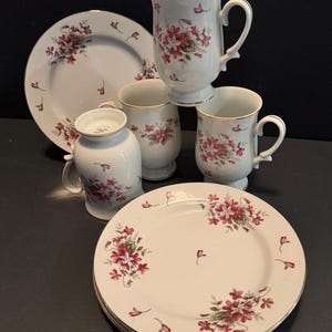 May include: A white porcelain tea set with a floral pattern. The set includes plates, mugs, and a small pitcher. The floral design features red flowers and green leaves, with gold trim. The set is arranged on a dark surface.