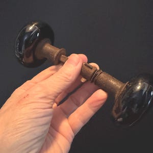 May include: Antique door knob with black, glossy ceramic knobs and a rusty metal rod. The door knob is held in a hand against a dark background. The door knob is a vintage item.