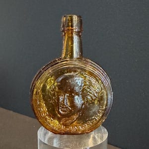 May include: Amber-colored glass bottle with a round body and a narrow neck. The front features a detailed relief of a person's face within a circular frame. The bottle is likely an antique or vintage collectible.