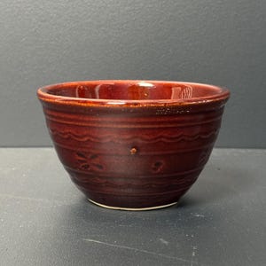 May include: A small, red ceramic bowl with a wavy rim and a decorative pattern. The bowl is glazed in a deep, rich red color.