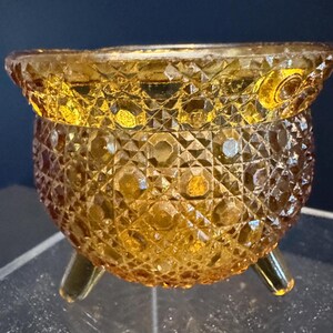May include: A vintage amber-colored glass bowl with a diamond-cut pattern. The bowl has a rounded body and sits on three small, curved legs. The rim of the bowl has a decorative edge. This decorative bowl is perfect for serving snacks or as a decorative piece.