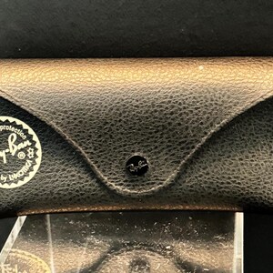 Pu&ograve; includere: Un astuccio nero in pelle per occhiali da sole Ray-Ban con superficie strutturata e parte superiore color oro. L'astuccio ha una chiusura a scatto e un adesivo bianco con il logo e il testo Ray-Ban.