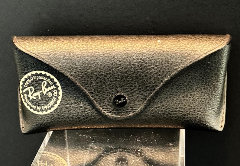 Pu&ograve; includere: Un astuccio nero in pelle per occhiali da sole Ray-Ban con superficie strutturata e parte superiore color oro. L'astuccio ha una chiusura a scatto e un adesivo bianco con il logo e il testo Ray-Ban.