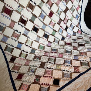 May include: A handmade quilt with a repeating pattern of white squares and brown rectangles. The squares are separated by a white fabric and the rectangles contain various floral and patterned fabrics. The quilt is trimmed with a dark blue border.