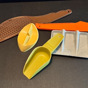 May include: A collection of vintage kitchen tools in various colors. Includes a brown grater, an orange knife, a yellow scoop, a yellow citrus juicer, and a gray tray. These tools are made of plastic and are arranged on a dark surface.