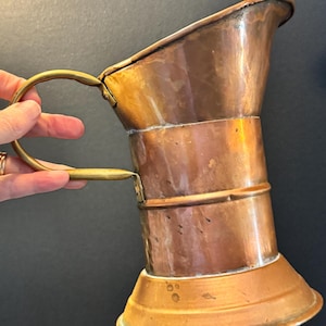 May include: A copper pitcher with a brass handle. The pitcher has a flared base and a cylindrical body with a spout. The handle is attached to the top and bottom of the pitcher. The copper has a warm, reddish-brown hue.