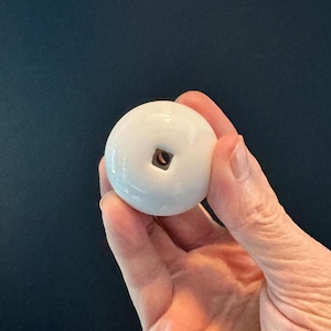 May include: A white, spherical ceramic knob with a square inset. The knob is held in a hand against a dark blue background. This decorative hardware is suitable for cabinets, drawers, or furniture.