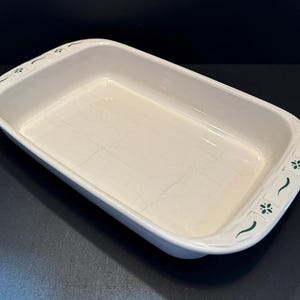 May include: A rectangular white ceramic baking dish with a glossy finish and visible fine crackle patterns throughout the interior. The handles feature a decorative green floral and curved line motif on each side of the rim of the dish.