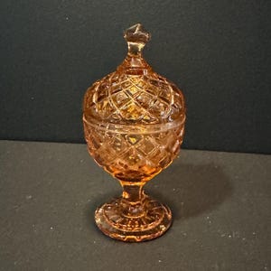 May include: Amber-colored, vintage glass candy dish with a lid. The dish has a diamond pattern and sits on a pedestal base. The lid is topped with a small, pointed finial. The glass has a warm, golden hue.