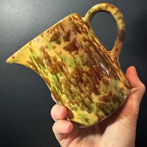 May include: A ceramic pitcher with a spout and handle. The pitcher has a speckled pattern in shades of yellow, brown, and green. The handle is arched, and the spout is pointed. The pitcher is held in a hand.