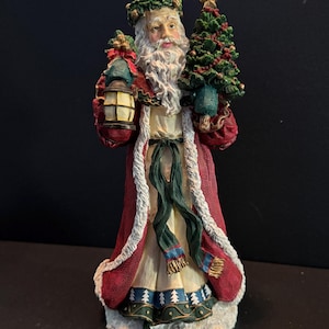 May include: A detailed figurine of Santa Claus holding a lantern and a Christmas tree. Santa wears a red coat with white trim, a green wreath, and a long white beard. The tree is decorated with red beads and a gold star.