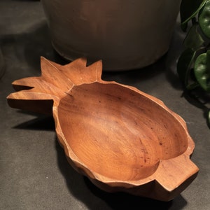 May include: A wooden pineapple-shaped bowl with a smooth, brown finish. The bowl is carved to resemble a pineapple, with a deep, rounded interior.