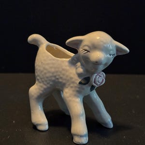 May include: A white ceramic lamb figurine with a small opening on its back. The lamb has a pink rose and green leaves on its chest. The figurine has a textured surface and is posed with its head tilted slightly.