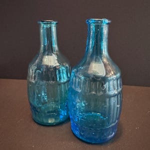 May include: Two blue glass bottles with a textured, barrel-like design. The bottles are a vibrant shade of blue and have a classic, vintage aesthetic. The bottles are empty and appear to be decorative or for storing liquids.