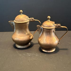 May include: Two antique copper teapots with brass accents. The teapots have a classic design with curved spouts, rounded bodies, and small handles. The lids are domed and topped with brass knobs. The copper has a warm, aged patina.