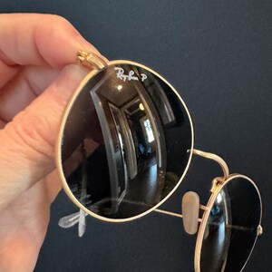 Pu&ograve; includere: Occhiali da sole con montatura dorata e lenti scure. Il logo Ray-Ban &egrave; visibile sulla lente superiore sinistra. Gli occhiali da sole hanno una classica forma rotonda.