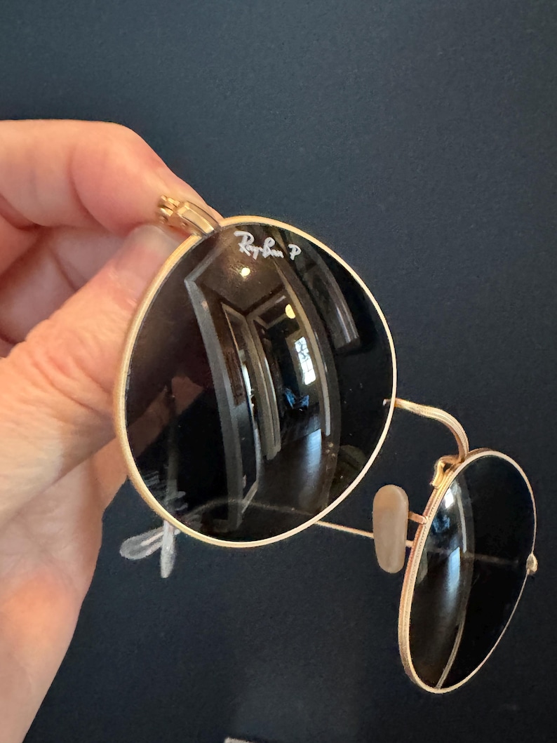 Pu&ograve; includere: Occhiali da sole con montatura dorata e lenti scure. Il logo Ray-Ban &egrave; visibile sulla lente superiore sinistra. Gli occhiali da sole hanno una classica forma rotonda.