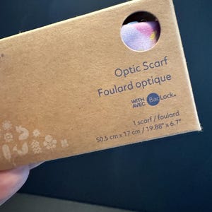 May include: A rectangular brown cardboard box containing an Optic Scarf. The packaging features the text Optic Scarf, Foulard optique, WITH AVEC BacLock, and 1 scarf / foulard. Dimensions are listed as 50.5 cm x 17 cm / 19.88 inches x 6.7 inches.