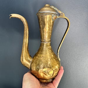 May include: A vintage brass pitcher with a long, curved spout and a handle. The pitcher has a hammered texture and a lid. The overall design is ornate and decorative, suitable for display or serving.