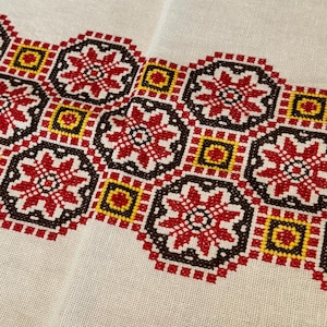 May include: A white fabric with a red, yellow, and brown embroidered border. The border features a repeating pattern of stylized flowers.
