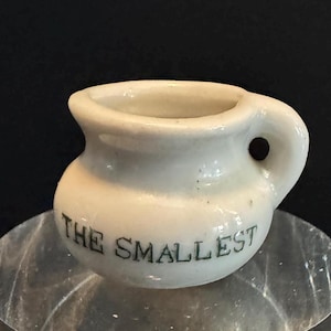 May include: A small, off-white ceramic pitcher with a rounded body and a handle. The words "THE SMALLEST" are printed in black on the front. The pitcher is likely a decorative item or a miniature collectible.