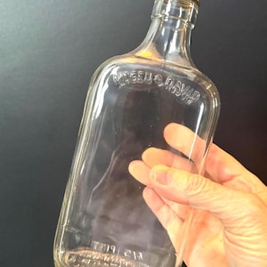 May include: A clear glass bottle with a white metal cap. The bottle has a rectangular shape with rounded edges and the embossed text "MOGEN & DAVID" and "ONE PINT". The bottle is held by a hand.