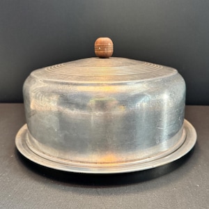 May include: A round, brushed aluminum food cover with a domed shape and a small, spherical wooden knob handle at the top. The cover features concentric circular grooves etched into the top surface and rests on a matching circular metal tray base.