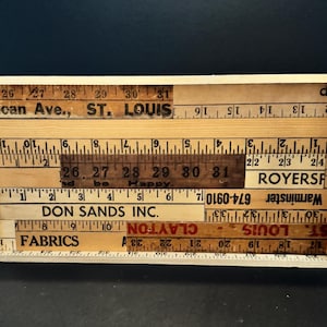 May include: A rectangular wooden art piece composed of vintage measuring tapes. The tapes, in various shades of brown, are arranged in a mosaic pattern. Text fragments like "St. Louis" and "Fabrics" are visible. The piece is framed in a light-colored wood.