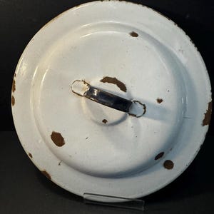 May include: A round, white enameled metal lid featuring a dark blue handle centered on top. The surface shows visible signs of wear, including several brown rust spots and chipped enamel along the edges and around the handle attachment points.