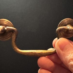 May include: A brass-toned metal drawer pull featuring a curved, swinging handle attached to two circular backplates. The hardware has a weathered, aged finish with visible texture and rounded details on the mounting points and the central grip.